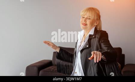 Ältere Frau mit Fransenlederjacke und weißem Hemd, glücklich tanzt mit geschlossenen Augen und lächelnd, bringt aktives Altern, Freude und Freiheit zum Ausdruck Stockfoto