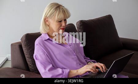 Ältere Frau mit blondem Haar, die ein lila Hemd trägt, sitzt bequem auf einem braunen Sofa, tippt aktiv auf einem Laptop und nimmt an Online-Aktivitäten Teil Stockfoto