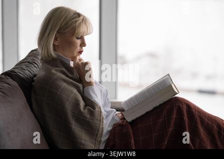 Seniorin in eine warme Decke auf einem Sofa gewickelt, liest und reflektiert an einem Fenster in einem gemütlichen Wohnzimmer, genießt friedliche, nachdenkliche Freizeit Stockfoto