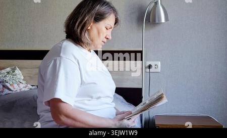 Seniorin genießt ruhige Zeit beim Lesen eines Buches, während sie auf ihrem Bett sitzt und einen Moment der Entspannung und Einsamkeit in ihrem komfortablen Zuhause findet Stockfoto
