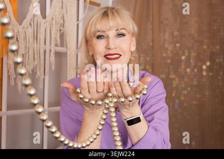 Die blonde Seniorin lächelt selbstbewusst, während sie eine Schnur aus silbernen dekorativen Perlen hält, während sie ein lila Hemd und roten Lippenstift in einer Halle trägt Stockfoto