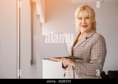 Geschäftsführerin steht in einem modernen Büro, hält selbstbewusst einen Laptop und lächelt in die Kamera, verkörpert Erfahrung und Digital Stockfoto