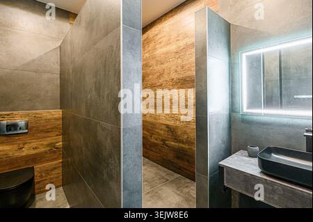 Das moderne, minimalistische Badezimmer ist mit grauen Fliesen, Holzakzenten, einer begehbaren Dusche und einem eleganten schwarzen Waschbecken und einer Toilette ausgestattet. Die sauberen Leitungen und Industrie Stockfoto