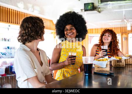 Multiethnische Gruppe junger glücklicher Freunde, die sich im Vintage Diner Burger House Restaurant zusammenschließen - drei lächelnde multiethnische Freundinnen genießen Drinks und Stockfoto