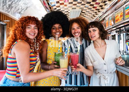 Multiethnische Gruppe junger glücklicher Freunde, die sich im Vintage Diner Burger House Restaurant zusammenschließen - verschiedene Freunde lächeln und bunten Milchshakes in A toasten Stockfoto