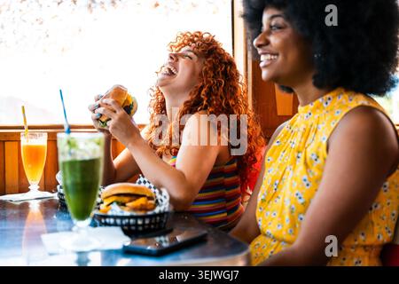 Multiethnische Gruppe junger glücklicher Freunde, die sich im Vintage Diner Burger House Restaurant zusammenschließen - multiethnische junge Frauen lachen und genießen Burger und Stockfoto