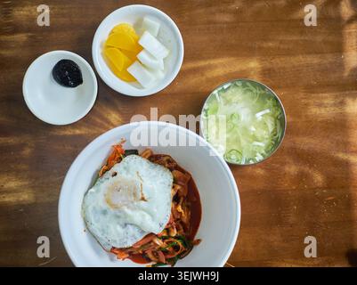 Dieses Bildset zeigt Junghwa Bibimbap mit scharfem gemischtem Reis mit Meeresfrüchten, einem Spiegelei und traditionellen Beilagen auf einem Holztisch Stockfoto