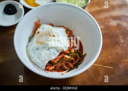 Dieses Bildset zeigt Junghwa Bibimbap mit scharfem gemischtem Reis mit Meeresfrüchten, einem Spiegelei und traditionellen Beilagen auf einem Holztisch Stockfoto