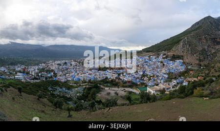 Das Panorama der Bergstadt in Chefchaouen zeigt weiße und blaue Gebäude, die sich über ein Tal unter zerklüfteten Hügeln unter bewölktem Winterlicht verteilen. Stockfoto