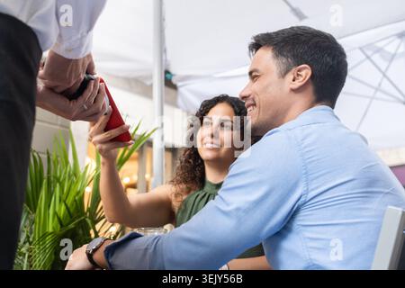 Glückliches junges Paar, das auf einer Terrasse im Freien sitzt und kontaktlos per Handy bezahlt. Der Kellner hält das Zahlungsterminal als Frau Stockfoto
