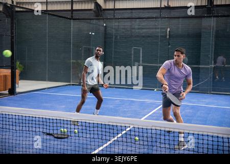 Verschiedene männliche Partner in Sportbekleidung, die sich auf dem blauen Padel-Platz tummeln und mit schwarzem Schläger Ball schlagen Stockfoto