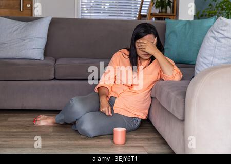 Reife Frau sitzt auf Hartholzboden mit grauem Sofa bedeckt Gesicht, Korallenhemd graue Hose, Becher Stockfoto