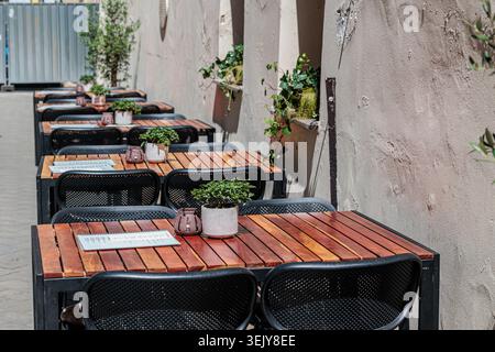 Leere Terrasse im Freien mit Holztischen, schwarzen Metallstühlen, kleinen Topfpflanzen und Menüs, die eine ruhige, sonnendurchflutete urbane Speiseszene schaffen Stockfoto
