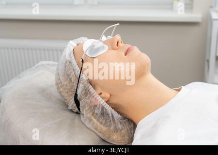 Entspannte Frau, die professionelle Gesichtsbehandlung mit verjüngender Gelmaske, Augenschutz und Einweg-Haarkappe in der Schönheitsklinik erhält Stockfoto