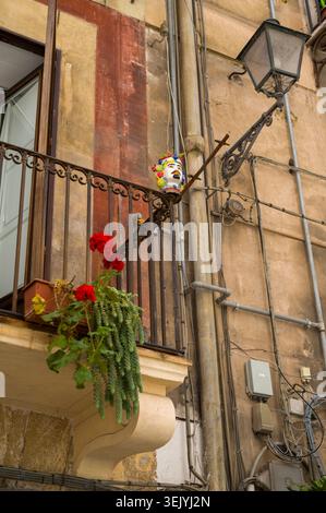 Auf einem rustikalen Eisenbalkon neben roten Geranien befindet sich ein klassischer sizilianischer Keramiktopf mit Moorkopf, der ein traditionelles Stück regionaler Folklore zeigt Stockfoto