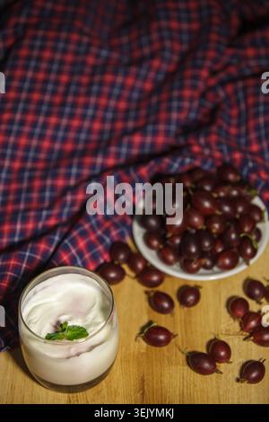 Frische Stachelbeeren und cremiger Joghurt im Glas auf einem Holztisch mit Minzblättern und kariertem Stoff. Rustikales hausgemachtes Frühstück oder gesundes Desse Stockfoto