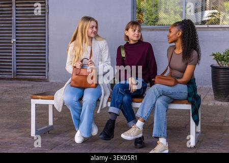 Verschiedene Freundinnen sitzen auf Holzbank am Fenster mit Handtasche und Rucksack Stockfoto