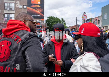 Drei Personen in roter und schwarzer Kleidung stehen eng zusammen, und eine Person auf einem Mobiltelefon. Der Hintergrund zeigt eine urbane Straßenszene mit Gebäuden, einer Werbetafel und anderen Personen. Stockfoto