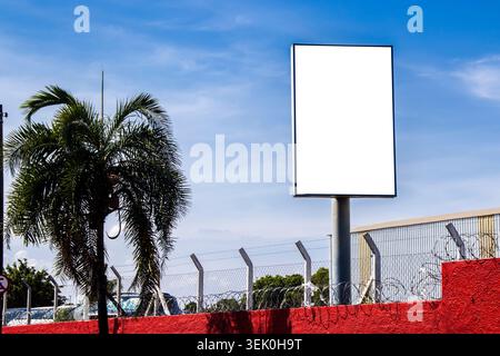 Vertikale leere weiße Werbetafel für Werbemockup hinter einer rot strukturierten Wand mit Stacheldrahtzaun. Industriegebäude und blauer Himmel B Stockfoto