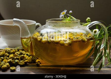 Kamille und Chrysanthemen Kräutertee in Glas-Teekanne mit getrockneten Blumen auf rustikalem Holztisch. Relaxationskonzept, traditionelle Kräutermedizin Stockfoto