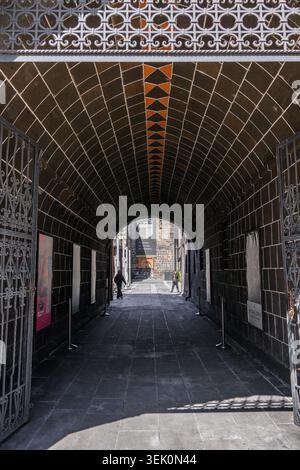 Gewölbter Gang mit komplizierten Metallarbeiten und gekachelter Decke. Sonnenlicht strömt durch einen offenen Bogen am Ende Der Bürgersteig ist dunkel und glatt mit heller Re Stockfoto