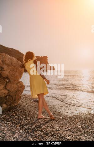Sonnenuntergang Frau Strand Frau in einem gelben Kleid lehnt sich auf einem Küstenfelsen bei Sonnenuntergang und beobachtet Ozeanwellen mit Kopierraum Stockfoto