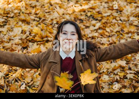 Fröhliche Erwachsene Frau liegt in einem Haufen Herbstlaub mit ausgestreckten Armen. Authentisches fröhliches Frauenporträt im Freien, fröhlicher Lebensstil und positiv Stockfoto