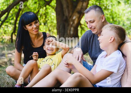 Mutter, Vater und zwei Söhne sitzen auf Gras unter einem großen Baum und lachen zusammen. Fröhliche Familie, die in einem Sommerpark eine schöne Zeit im Freien genießt. Stockfoto