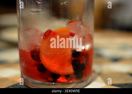 Nahaufnahme eines erfrischenden Getränks mit einer Scheibe Orange und gemischten Beeren in einem Glas mit Kondenswasser auf der Oberfläche, gegen ein sanft fokussiertes. Stockfoto