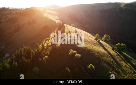 Atemberaubender Sonnenuntergang über hügeliger Landschaft. Die Sonne wirft warmes, goldenes Licht über Hügel und Täler und hebt das üppige Grün hervor. Die ruhige Szene, eingerahmt von fernen Bergen. Luftaufnahme Stockfoto
