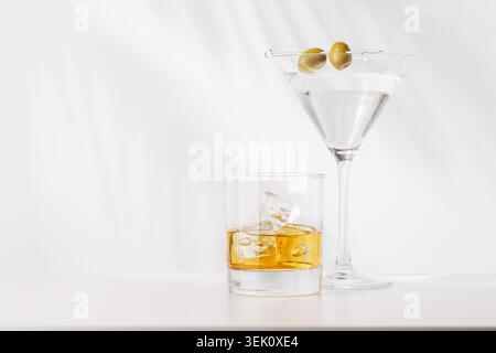 Elegante Komposition mit Whiskey auf den Felsen und Martini mit Oliven in Gläsern auf weißem Hintergrund. Klassische alkoholische Getränke mit Kopierraum Stockfoto
