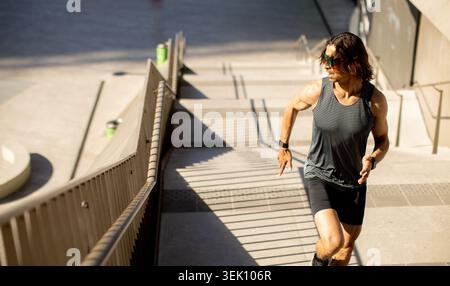 Eine Person fährt eine Treppe in einem Stadtpark hinauf. Die Sonne erzeugt Schatten auf den Stufen. Der Läufer ist fokussiert und trägt Sportausrüstung. Grelles Licht Stockfoto