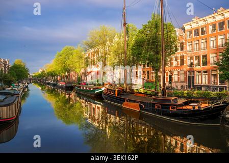 Traditionelle holländische Boote und historische Backsteinhäuser auf einem ruhigen Amsterdamer Kanal bei Sonnenaufgang, keine Menschen, Niederlande Stockfoto