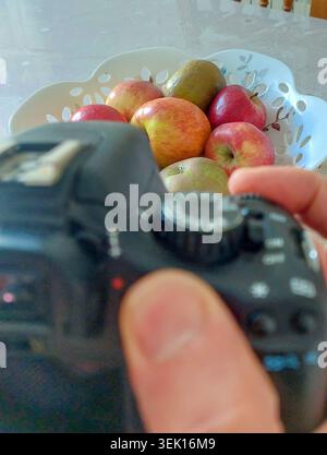 POV-Nahaufnahme einer Hand, die eine Digitalkamera hält, um eine Stillleben-Szene einer dekorativen Schüssel mit Bio-Äpfeln und Birnen festzuhalten, die Ho hervorhebt Stockfoto