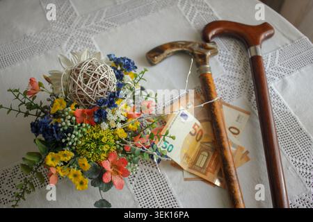 Eine konzeptionelle Hochwinkelaufnahme von zwei hölzernen Wanderstöcken, die über 50 Euro-Banknoten neben einem farbenfrohen Blumenarrangement auf einer weißen Tischdecke platziert haben Stockfoto
