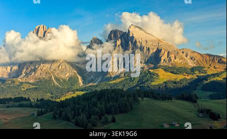 Compatsch, Italien - 30.09.2021: Zerklüftete Dolomitenberge, die sich durch Nebel über sanften grünen Hügeln und dunkle Kiefernwälder unter klarem blauem Himmel erheben Stockfoto