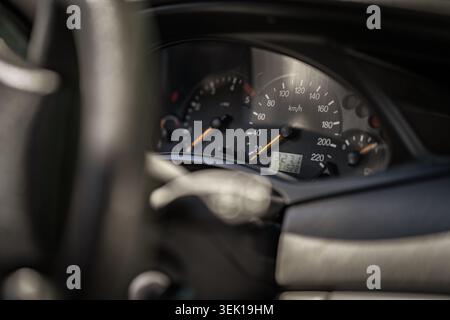 Armaturenbrett für das Fahrzeug mit einem analogen Tachometer, der die Geschwindigkeit in km/h anzeigt, einem Kilometerzähler, der die digitale Kilometerleistung anzeigt, sowie anderen Anzeigen und einer verschwommenen Straße Stockfoto