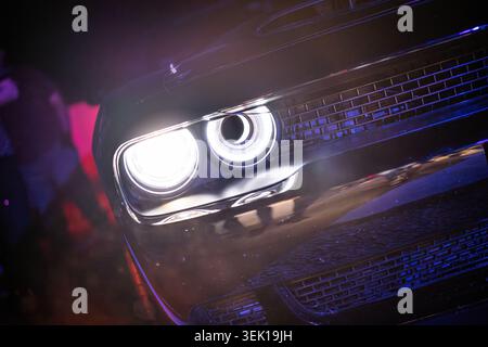 Dunkler Sportwagen mit beleuchteten LED-Scheinwerfern und Kühlergrill, der bei Nacht violettes und blaues Licht reflektiert und Fahrzeugdesign und fortschrittliche Technologie in Szene setzt Stockfoto