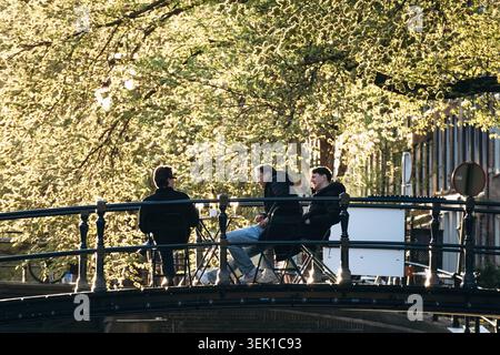 Amsterdam, Niederlande - 18. April 2026: Freunde, die sich auf einer Kanalbrücke in der warmen Abendsonne unter den Frühlingsbäumen im Zentrum von Amsterdam entspannen Stockfoto