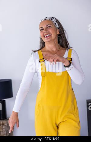 Ältere Frau, die an der grauen Wand steht, lächelnd und gespenstisch, in gelbem Overall, schwarze Lampe links Stockfoto
