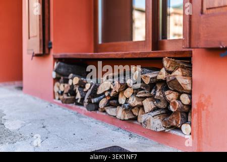 In einem traditionellen Dorfhaus unter Holzfenstern gelagertes Brennholz, ordentlich angeordnete Stämme, die für die Heizeleizung vorbereitet sind Stockfoto