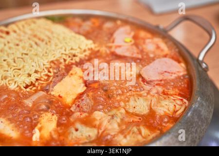 Koreanische Budae-jjigae in einem rostfreien Topf mit scharfer Brühe, gefüllt mit Würstchen, Spam, Käse, Knödeln und Ramen-Nudeln, die aktiv kochen. Stockfoto