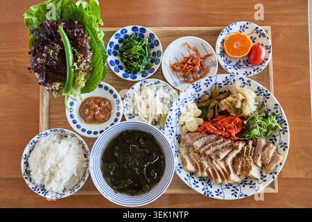 Ein traditionelles koreanisches Baekban-Menü mit gekochtem Schweinefleisch, Reis, Algensuppe, frischem Gemüse und verschiedenen Beilagen, die ordentlich auf einem Holztris angeordnet sind Stockfoto