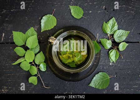 Über dem Blick auf Birkenbaum Blatt Tee in einer Glasschale, Betula medizinischen Kräutertee in einer klaren Tasse. Schwarzer Hintergrund aus Holzbrett, Blätter und Ast. Stockfoto