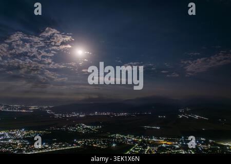 Die Luftaufnahme zeigt eine weitläufige Siedlung, die von einem hellen Vollmond beleuchtet wird. Die ruhige nächtliche Landschaft bietet leuchtende Lichter von Häusern und ferne Hügel unter einem tiefblauen Himmel. Stockfoto