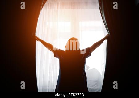 Frau, die Vorhänge vor einem hellen Fenster mit sanfter Hintergrundbeleuchtung öffnet Stockfoto