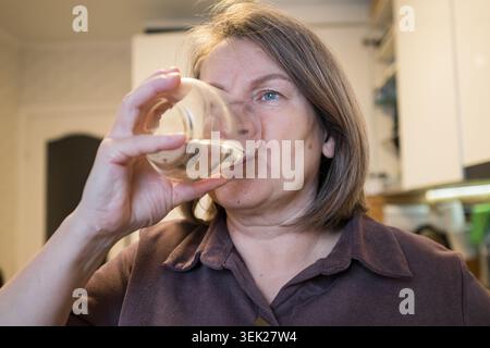 Reife Frau trinkt klares frisches Wasser aus einem Glas, versorgt und erfrischt sich in einer zwanglosen Innenküche, fördert Wohlbefinden und ein Stockfoto