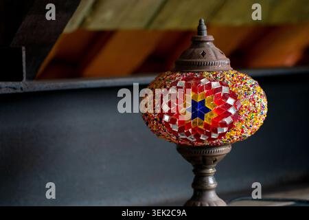 Eine traditionelle handgefertigte türkische Mosaiklampe mit roten, gelben und blauen Glasfliesen in einem geometrischen Sternmotiv, die vor einer dunklen Architektur platziert ist Stockfoto