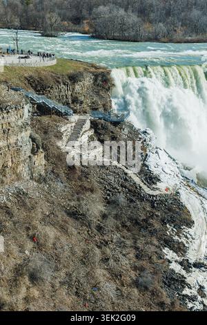 Touristen treffen sich am Rande der Niagarafälle, um den Blick auf das rauschende Wasser zu genießen. Das Gebiet zeigt auftauendes Eis und felsiges Gelände, wenn der Frühling eintrifft Stockfoto