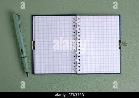 Offenes leeres Raster-Notizbuch liegt neben grünem Füllfederhalter auf grün strukturierter Lederoberfläche Stockfoto